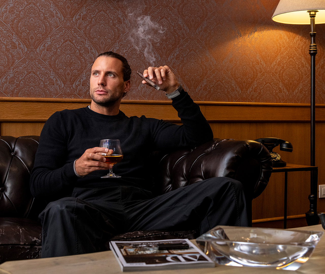 A guest relaxing in the cigar room, seated on a leather sofa with a cigar and a spirit