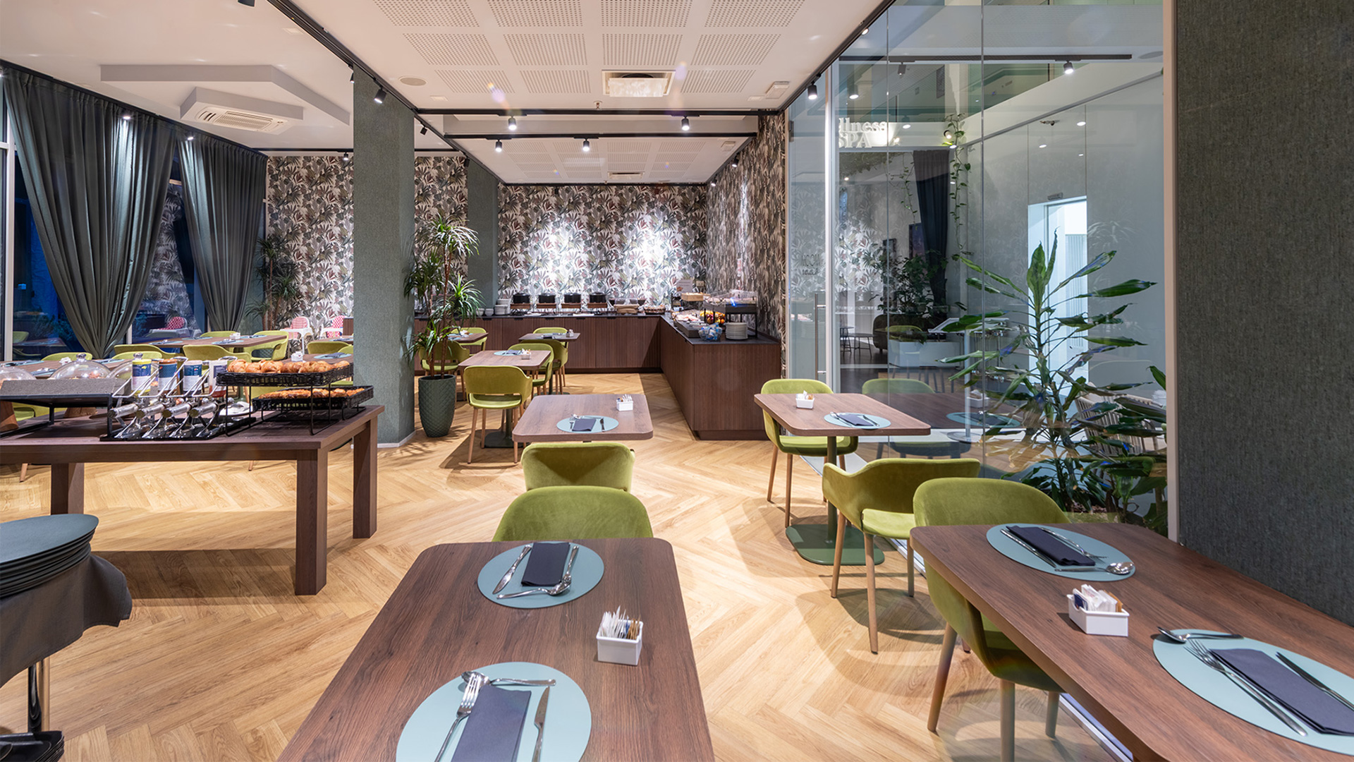 Sala ristorante luminosa con arredi moderni, sedie verde velluto, tavoli in legno e grandi vetrate con tende