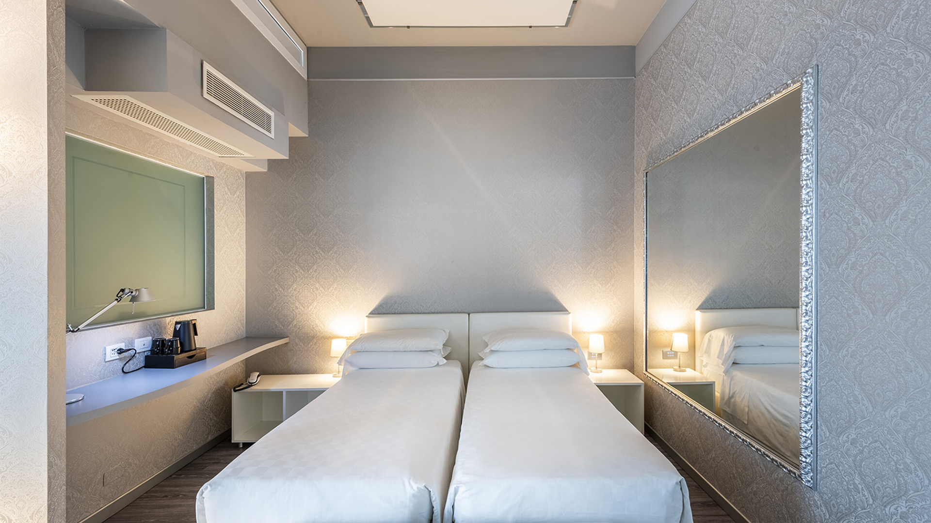 Deluxe Room with two single beds side-by-side, nightstands with lamps, large wall mirror with a silver frame, and a shelf with a kettle and coffee machine.