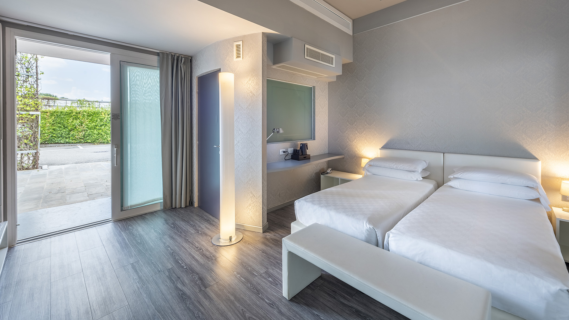 Deluxe Room with twin beds, bench at the foot of the bed, floating desk, floor lamp, and French door with direct outdoor access