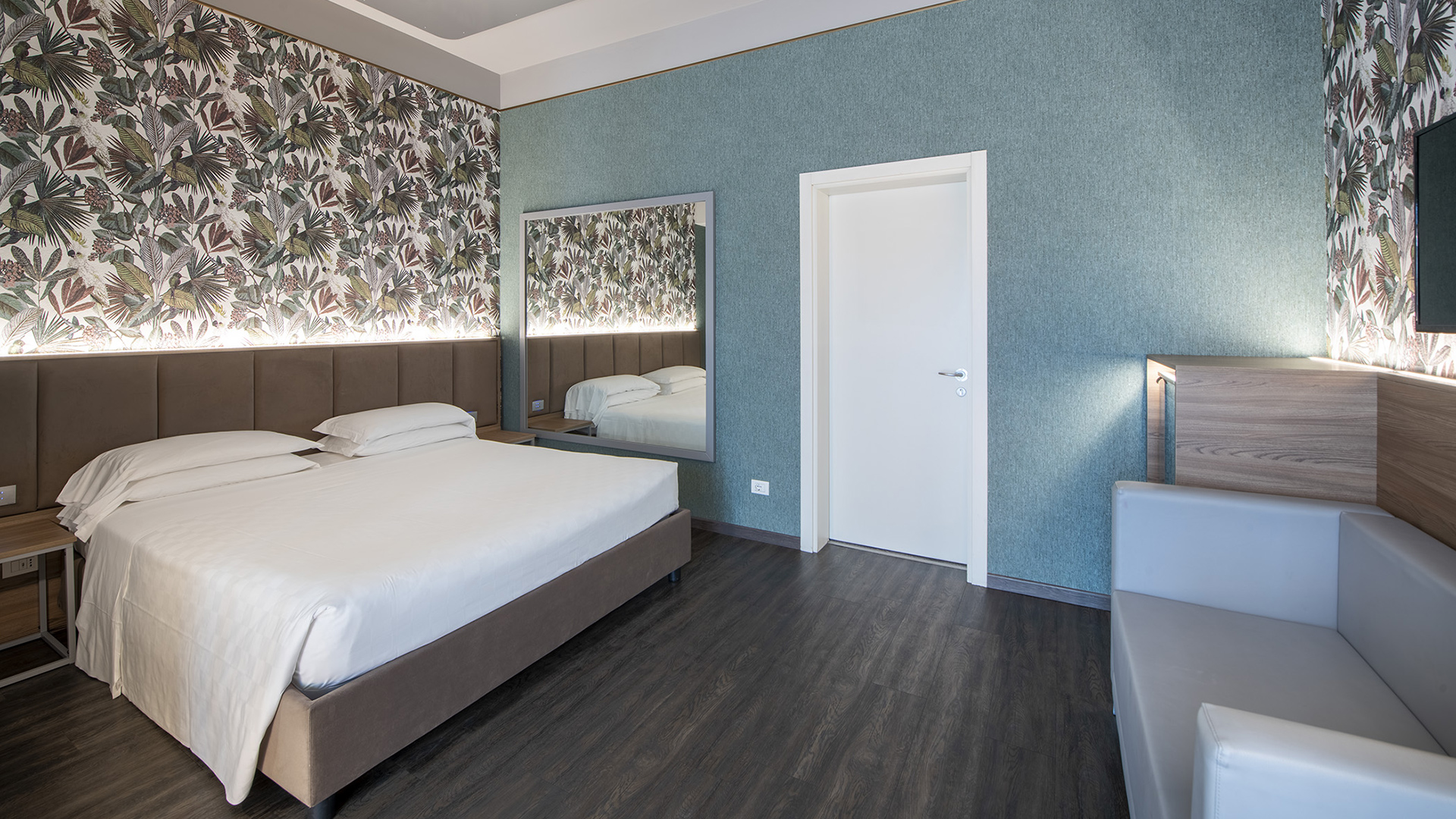 Deluxe Room with a double bed, a decorative wall featuring floral wallpaper, a large backlit mirror, wood-effect flooring, and a small side sofa.