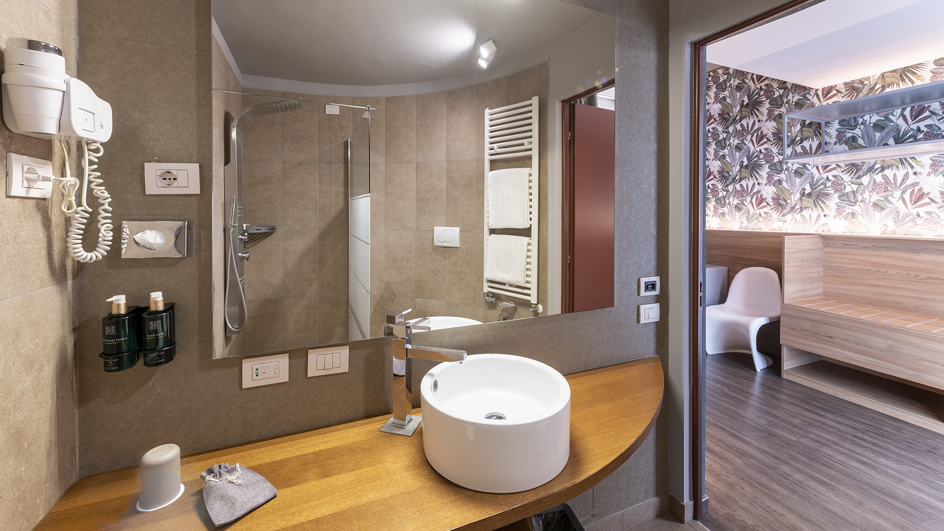 Modern bathroom with a round sink on a wooden countertop, large mirror, walk-in shower, and a view of the Deluxe room with floral wallpaper and wooden furniture.