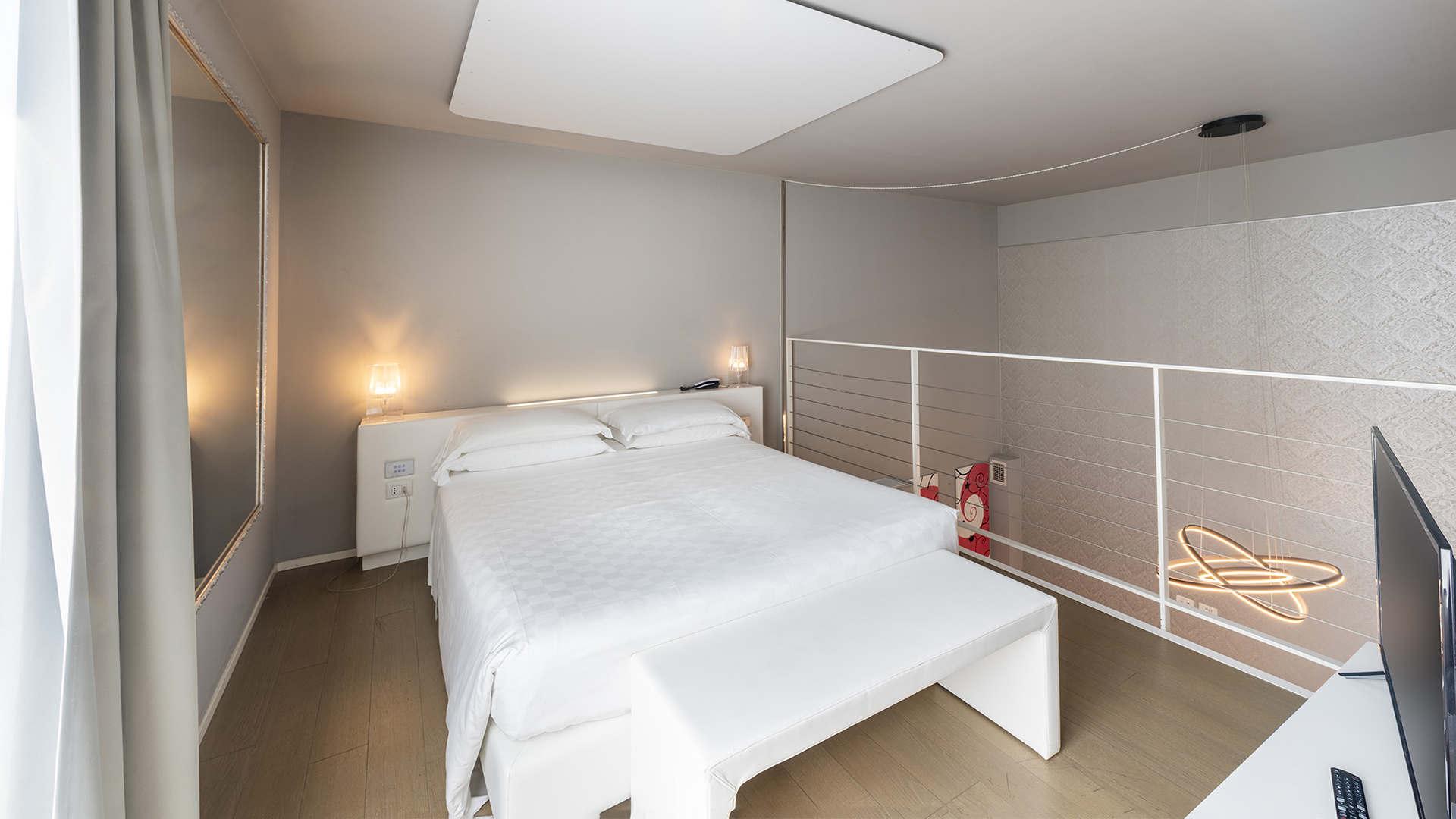 Junior Suite Deluxe su due livelli con letto matrimoniale, panca ai piedi del letto, lampade da comodino, soppalco con ringhiera bianca e lampada a sospensione di design