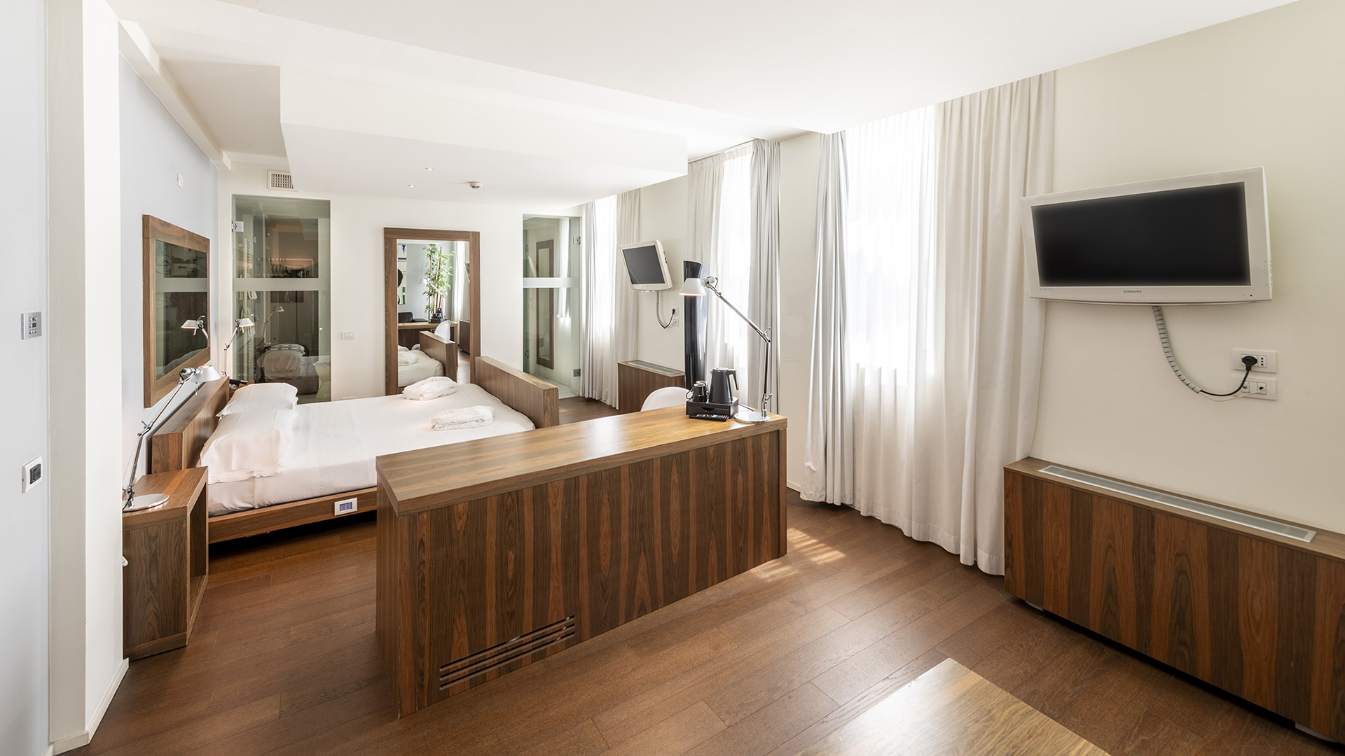 Superior Suite with a wooden double bed, partition cabinet with integrated desk, large wall mirror, wall-mounted TV, and spacious windows with white curtains