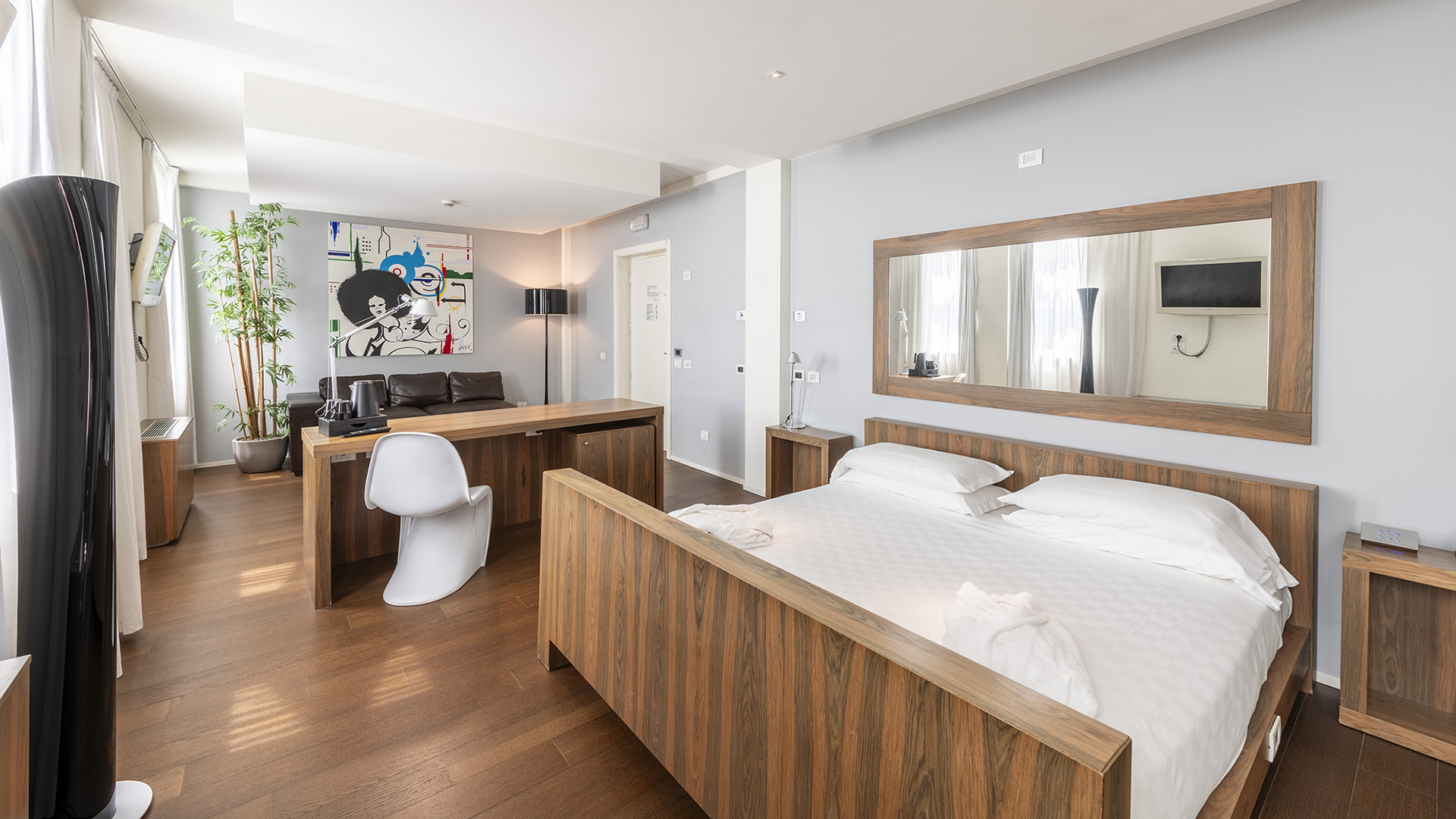 Superior Suite with a wooden double bed, central desk with a designer chair, living area with a leather sofa, large wall mirror, and large bright windows
