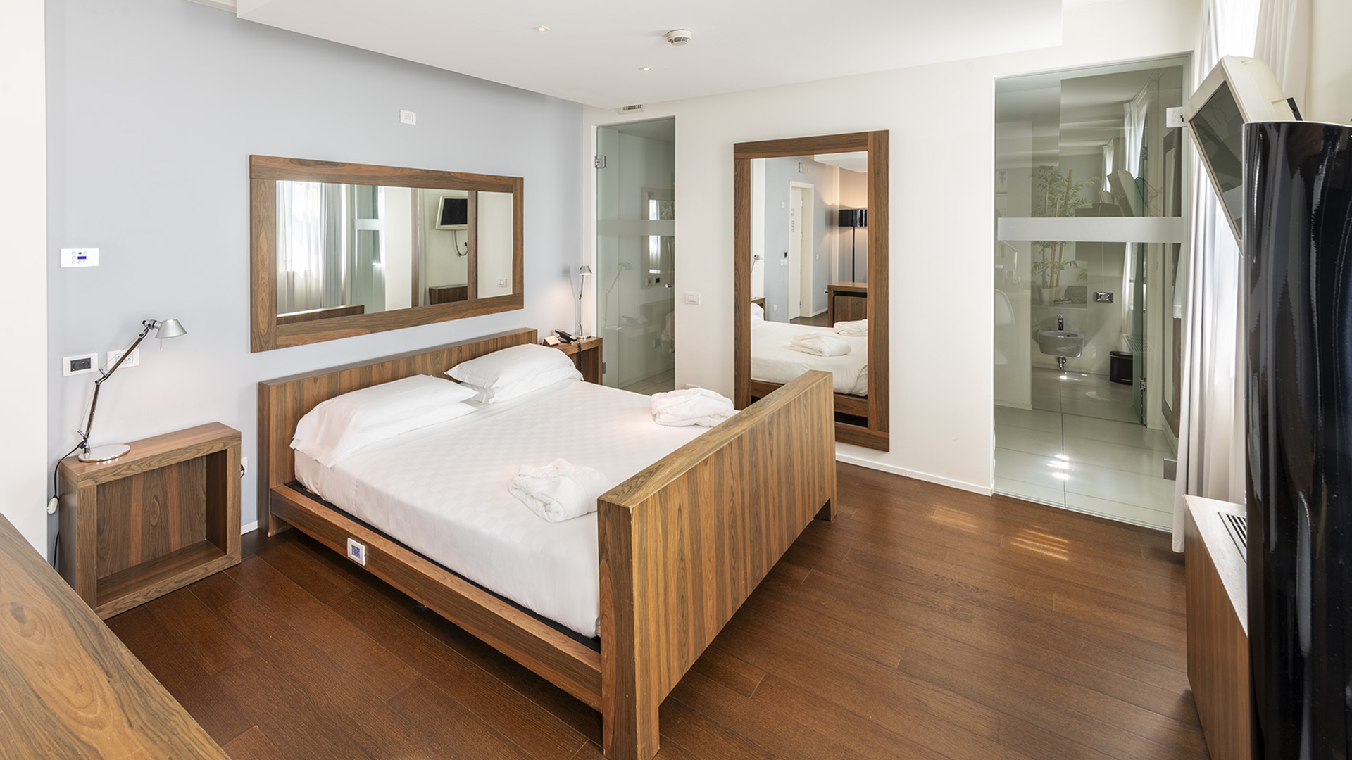 Superior Suite with a wooden double bed, matching nightstands, a large wall mirror, parquet flooring, and a bathroom with exposed glass walls.