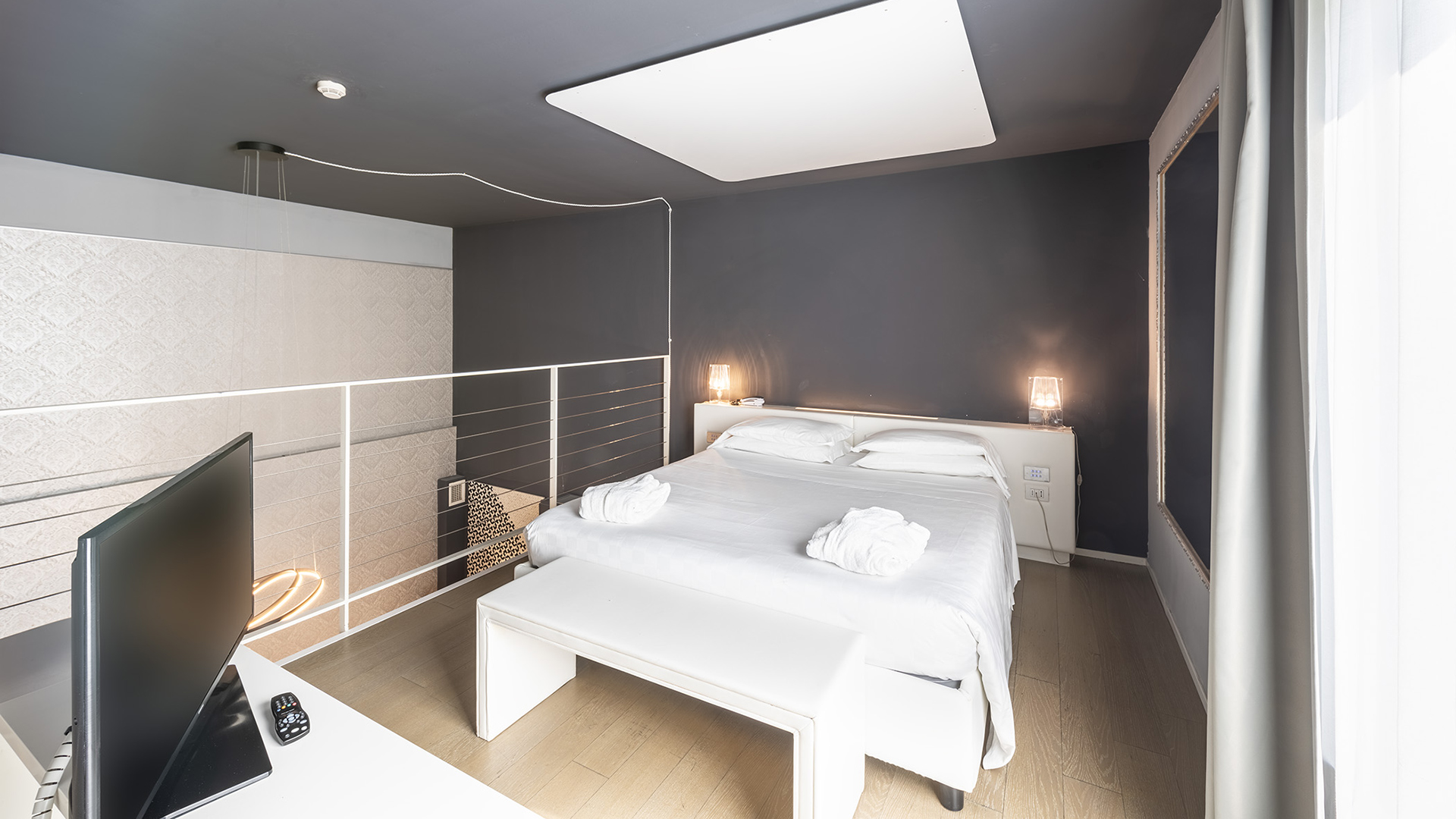 Elegant suite of the MO.OM Hotel, bright with a large window, central bed, dark walls, and design lamps.