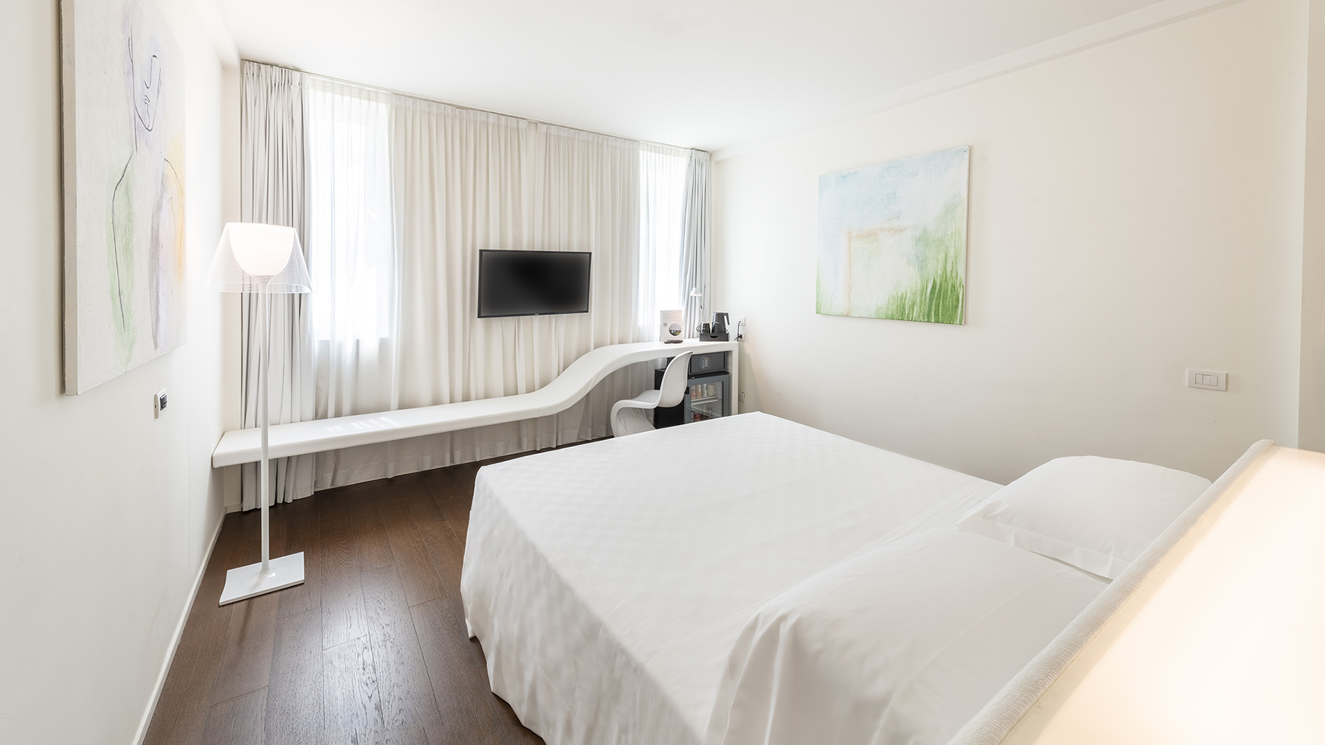 Superior Room with a double bed, a long white bench under the window with an integrated desk, a wall-mounted TV, and large bright curtains.