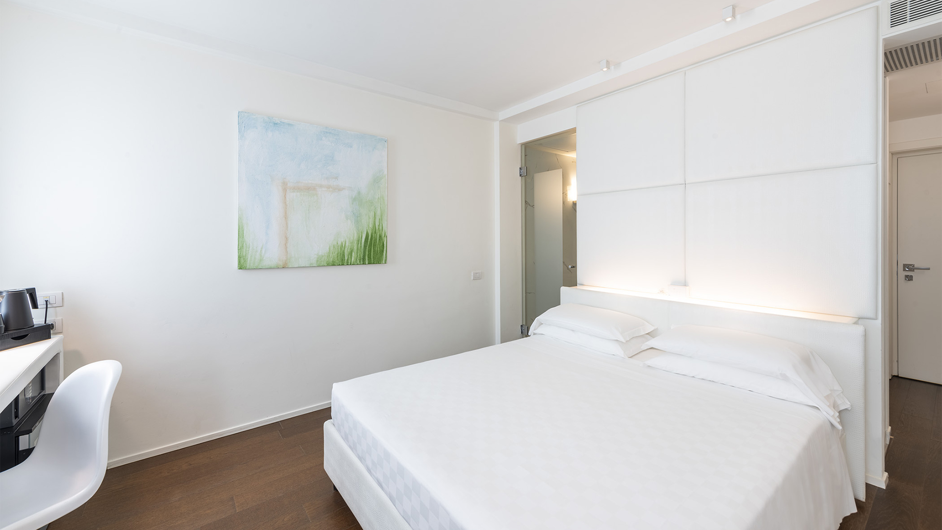 Superior Room with a double bed, white upholstered headboard with integrated lighting, side desk, and minimalist walls with an abstract painting