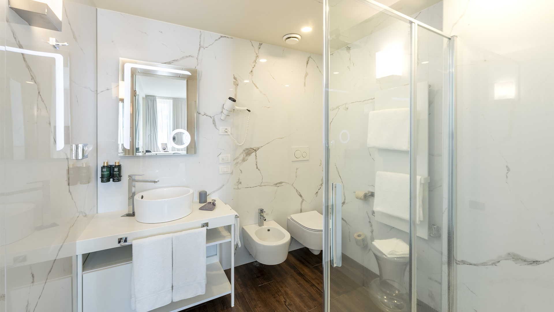 Modern bathroom with marble-effect tiles, countertop basin on a white vanity, illuminated mirror, glass shower, bidet, and wall-mounted toilet.